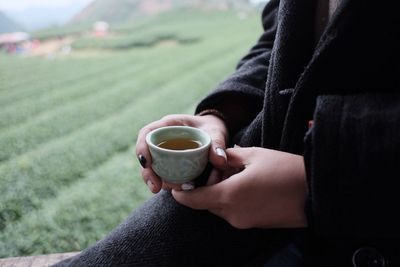 Midsection of woman holding coffee while sitting against field