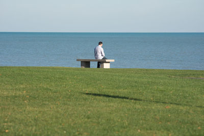 Rear view of man sitting on bench at field by sea against clear sky