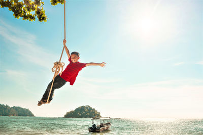 Boy swinging on rope swing over sea against sky during sunny day