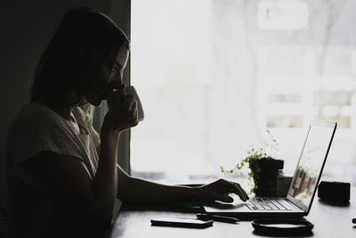 Side view of woman drinking coffee while using laptop against window