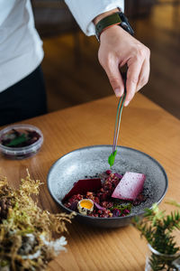 Cropped unrecognizable person preparing on a ceramic bowl with salad made with marinated herring and wild red lingonberries garnished with raw quail egg on wooden table