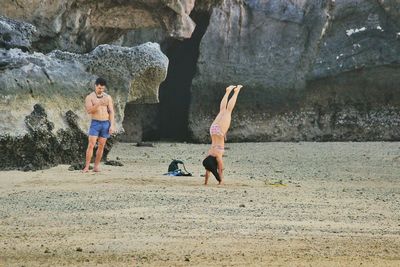 Shirtless man photographing woman doing handstand at beach against mountain