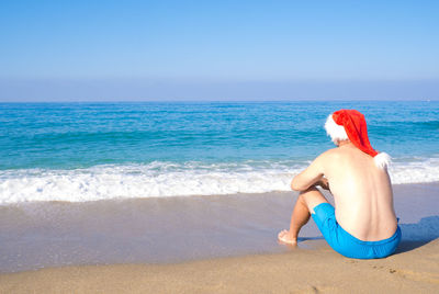 A man in a santa claus hat sits on the seashore, rear view. new year travel destinations