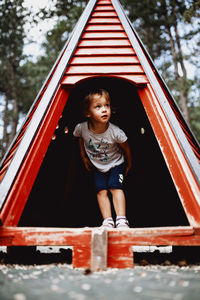 Child playing in a red woden tent at the natural kids playground surrounded with pine tree forest.