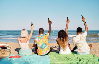 Cheerful multiethnic men and women raising bottles of beer and smiling while sitting on sand during picnic on beach in weekend