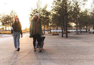 Mother and girl walking with luggage on footpath at sunset