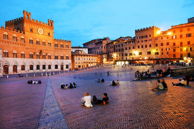 People relaxing by historic buildings at piazza del campo against sky during dusk