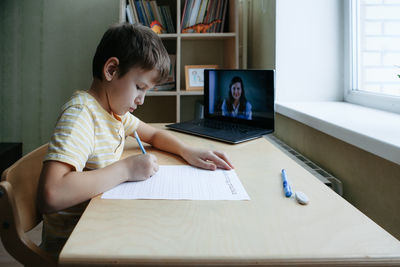 7 years old boy sitting by desk with laptop doing writing task during online lesson. side view