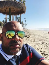 Portrait of bald man wearing sunglasses sitting on beach against clear sky