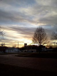 Bare trees by road against cloudy sky