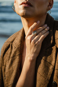 Crop ethnic man touching trendy jacket and looking at camera on blurred background of sea in nature