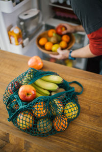 High angle of eco friendly cotton sack with ripe fruits placed on table in kitchen on background of crop person