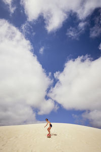 Girl sandboarding in desert against cloudy sky