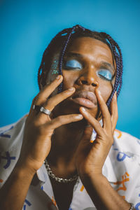 Serious african american male with braided hairstyle and bright makeup with closed eyes and hands on the face on blue background in studio