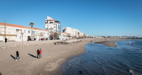 People on beach by buildings against clear blue sky