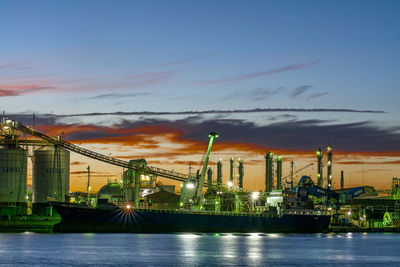 Illuminated factory by sea against sky during sunset