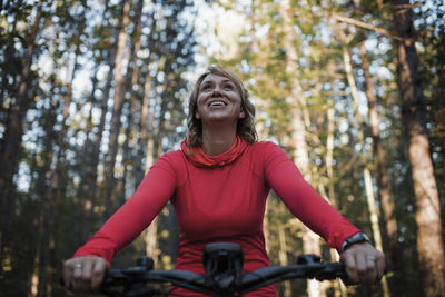 Low angle view of cheerful woman mountain biking against trees in forest