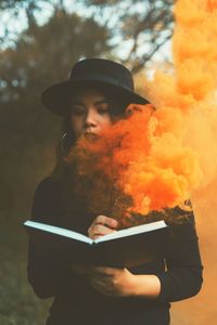 Young woman holding book while standing against trees in park