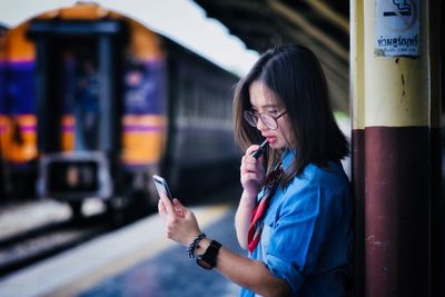 Side view of young woman applying lipstick while looking at mobile phone screen