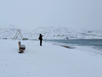 Man photographing at frozen lakeshore against clear sky during winter