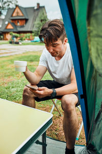 Young man drinking a coffee sitting at front of tent in the morning. teenager enjoying free time 