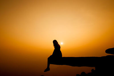 Silhouette woman sitting on cliff against sky during sunset