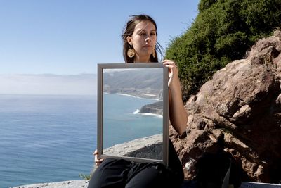 Portrait of young woman sitting on rock against sea