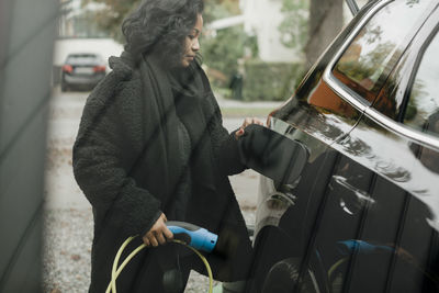 Mid adult woman with electric car at charging station seen through glass