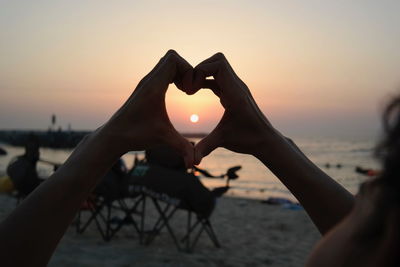 Cropped hands making heart shape at beach against sky during sunset