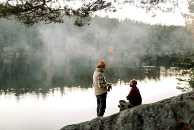 Male friends talking to each other on rock near lake during staycation