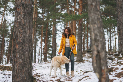 Young smiling woman in yellow jacket with big kind white dog labrador walking in winter forest