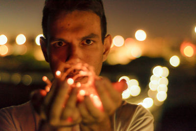 Close-up portrait of young man holding illuminated lighting equipment at night