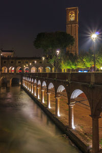 Illuminated bridge over river by buildings against sky at night