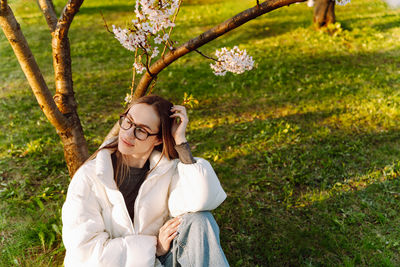 Beautiful young woman in glasses and white coat sits near blooming tree