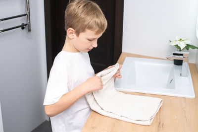The boy is standing in the bathroom near the washbasin and folds a towel