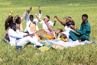 Cheerful black men and women in trendy clothes with fruits raising arms and smiling while sitting on blanket during picnic in grassy field