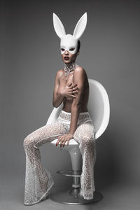 Portrait of young woman covering breast while sitting on chair against gray background