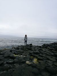 Rear view of woman standing on basalt columns by sea against sky at giant causeway