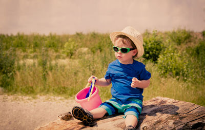 Cute boy in sunglasses and hat with bucket sitting against trees