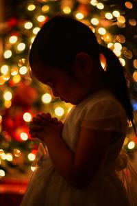 Side view of girl praying against illuminated lights