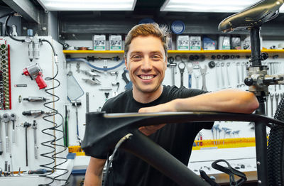 Happy male mechanic smiling and looking at camera while leaning on bike under repair against wall with tools in garage