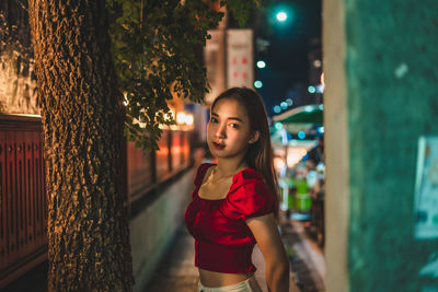 Portrait of young woman standing on sidewalk in city at night