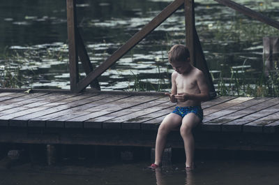 Shirtless boy sitting on pier over lake