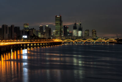 Illuminated buildings by river against sky at night
