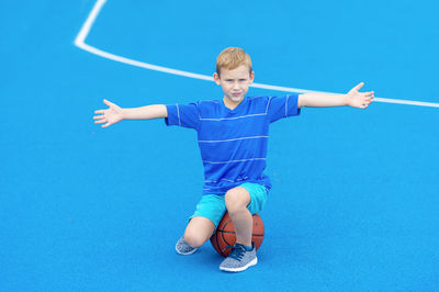 Portrait of boy with arms outstretched sitting on basketball at court