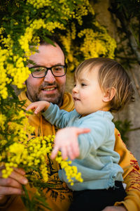 Little child sitting in arms of her father and playing with blooming mimosa plant nearby