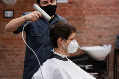 Barber in protective mask and latex glove styling adult man in a barbershop salon. new normal