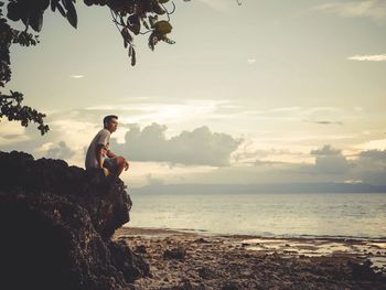 Man standing on rock at beach against sky during sunset