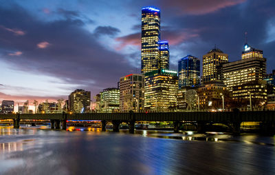 Illuminated modern buildings by river against sky at dusk