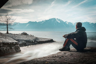 Rear view of man sitting on rock against mountains